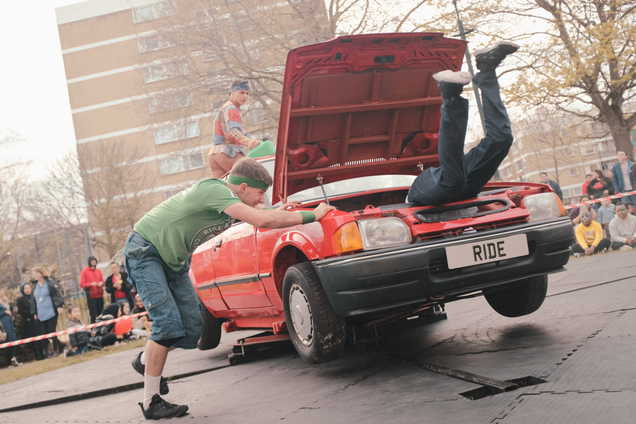 Three dancers perform on an old red Ford Orion car in the middle of a housing estate in Southampton.One dancer is pushing the car which is making it spin. The bonnet is popped up and the legs of another dancer are flailing out of the bonnet. There is an audience in the background watching on