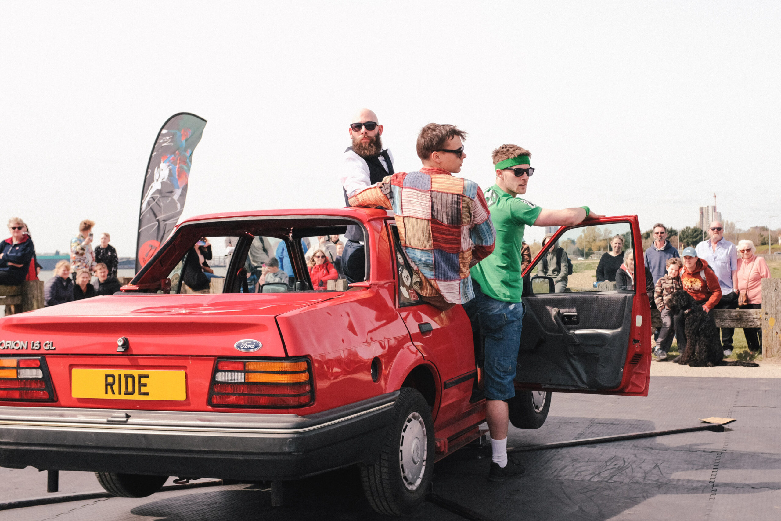 Three dancers perform on an old red Ford Orion car surrounded by an audience. They are leaning out the driver side window and door and are looking cool with some shades on