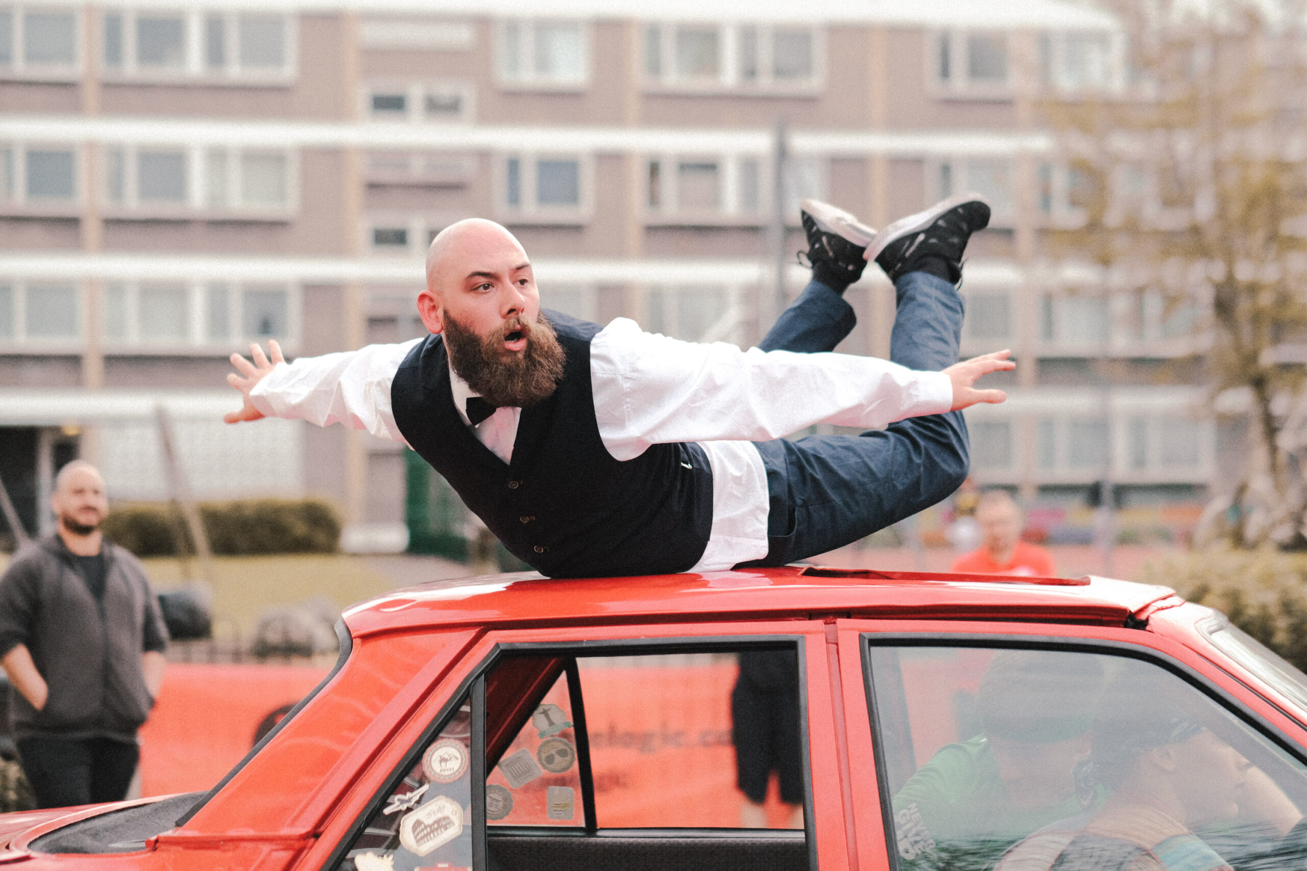A dancer spins on his stomach on top of a red car. He's dressed in a suit and his arms are outstretched and his feet are bent back into the air