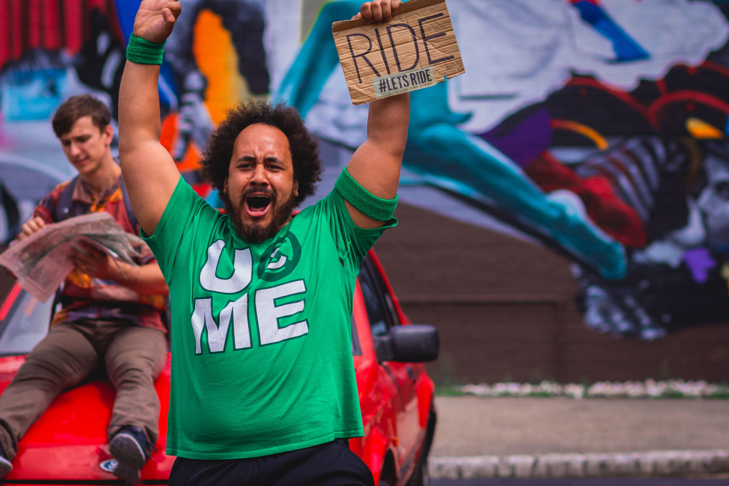 A man in a green tshirt stands with his arms in the air in front of a red car