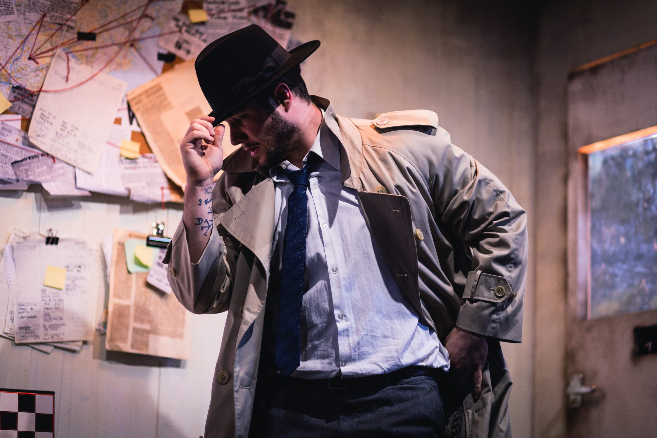 A dancer performs on a set which looks like a classic detective's office with papers on the wall and red string linking different bits of evidence. The dancer is wearing detective clothes - a black hat, long brown coat and shirt and tie