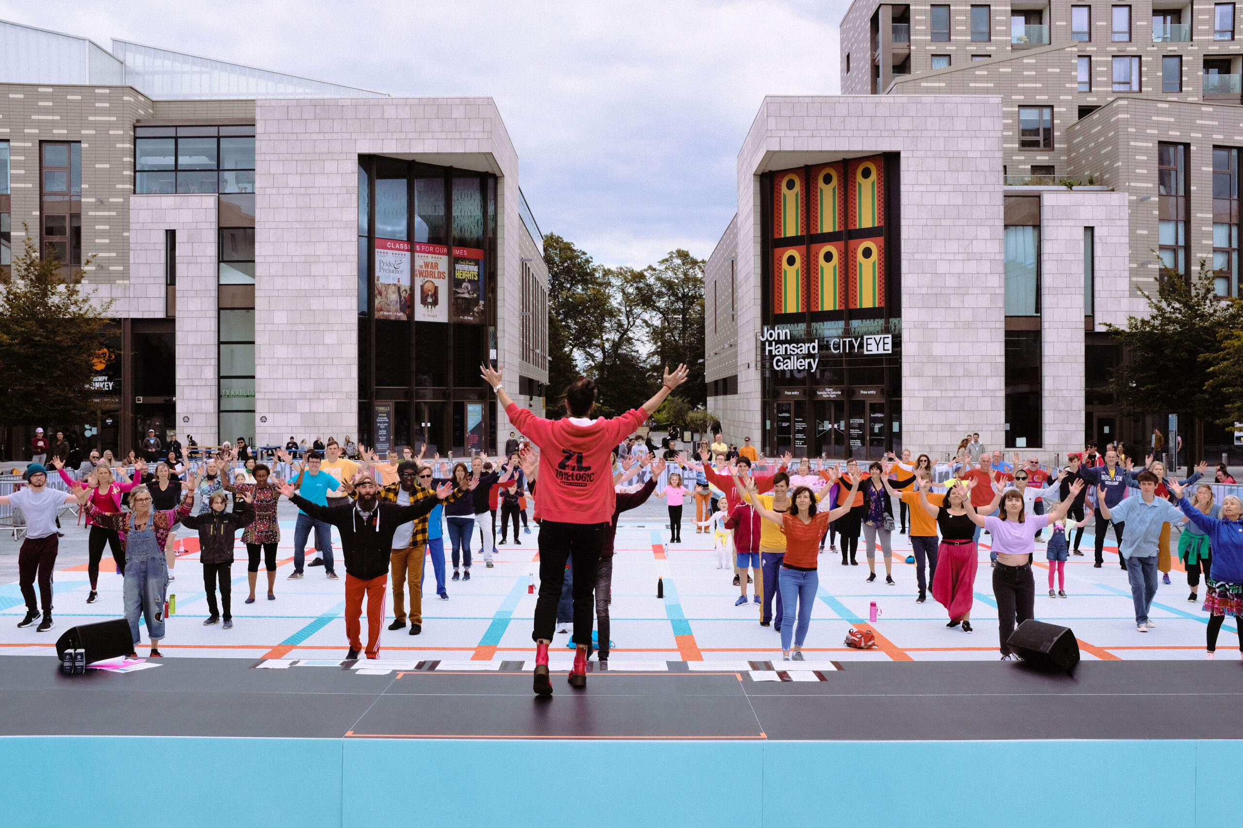 Participants performing in a socially distanced grid all hold their arms up in the air