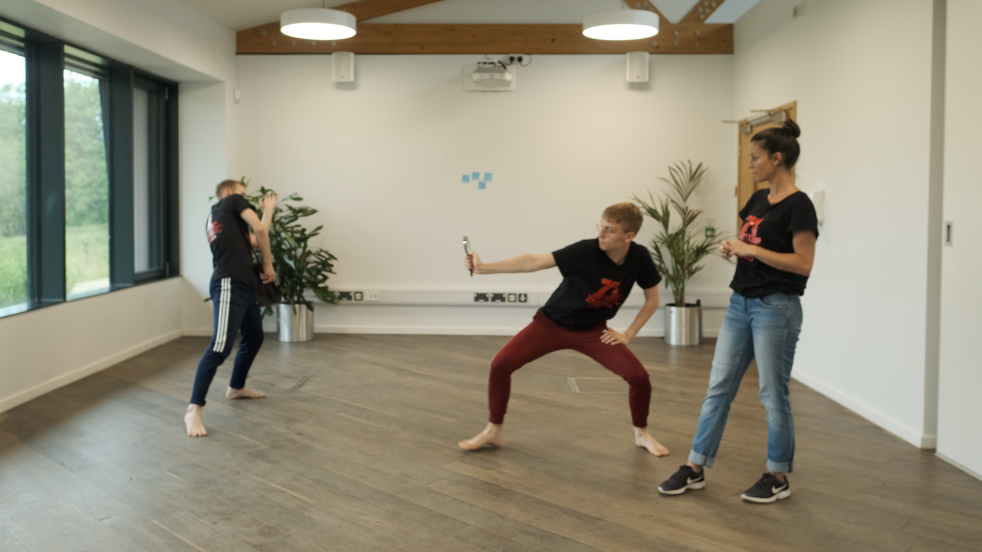 Two dancers and a choreographer are working together in a studio space. The dancers are moving and creating shapes with a prop in their hand