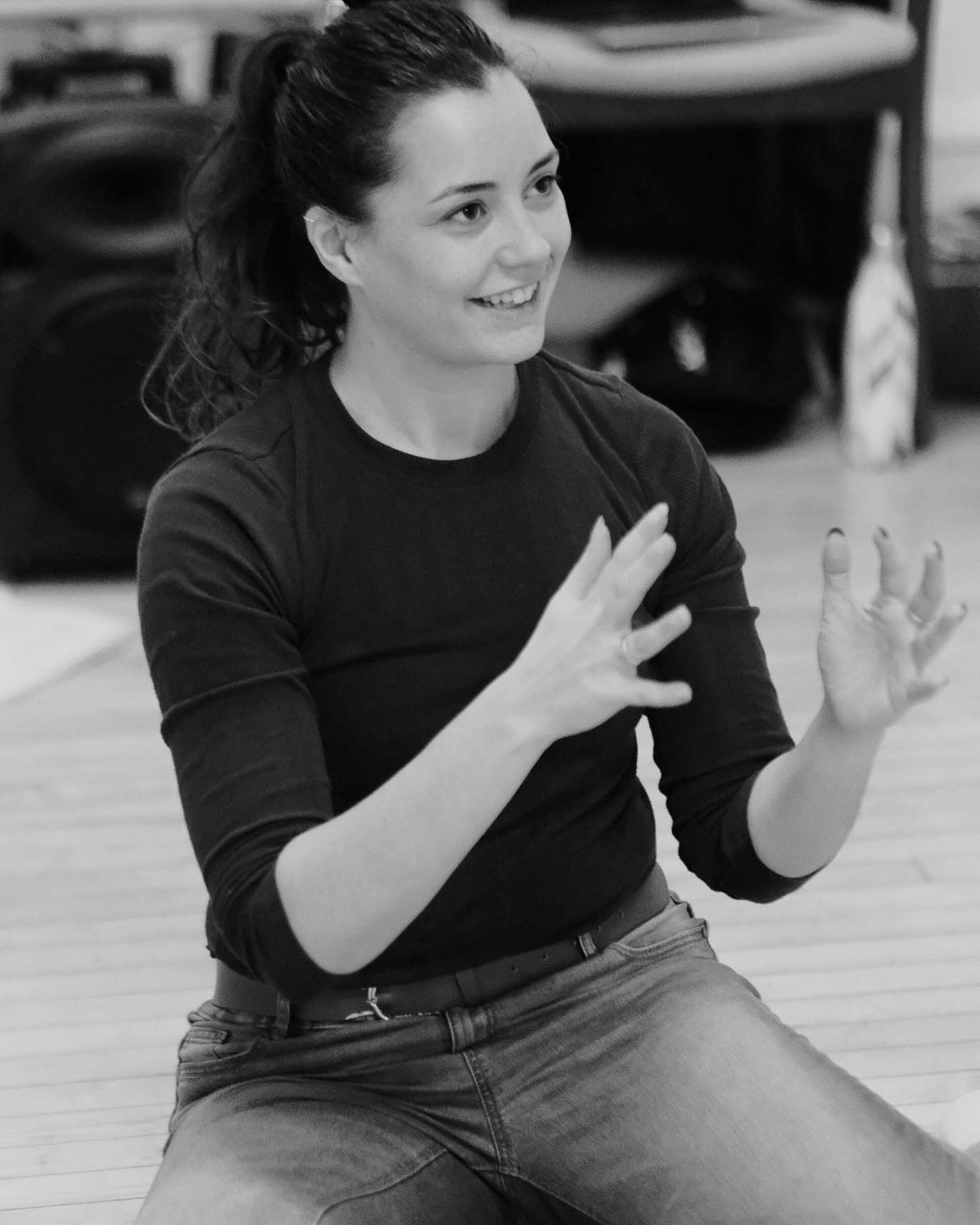 A picture of a young woman sat on the floor, smiling with her hands reaching out. It looks as though she is shaping something with her hands