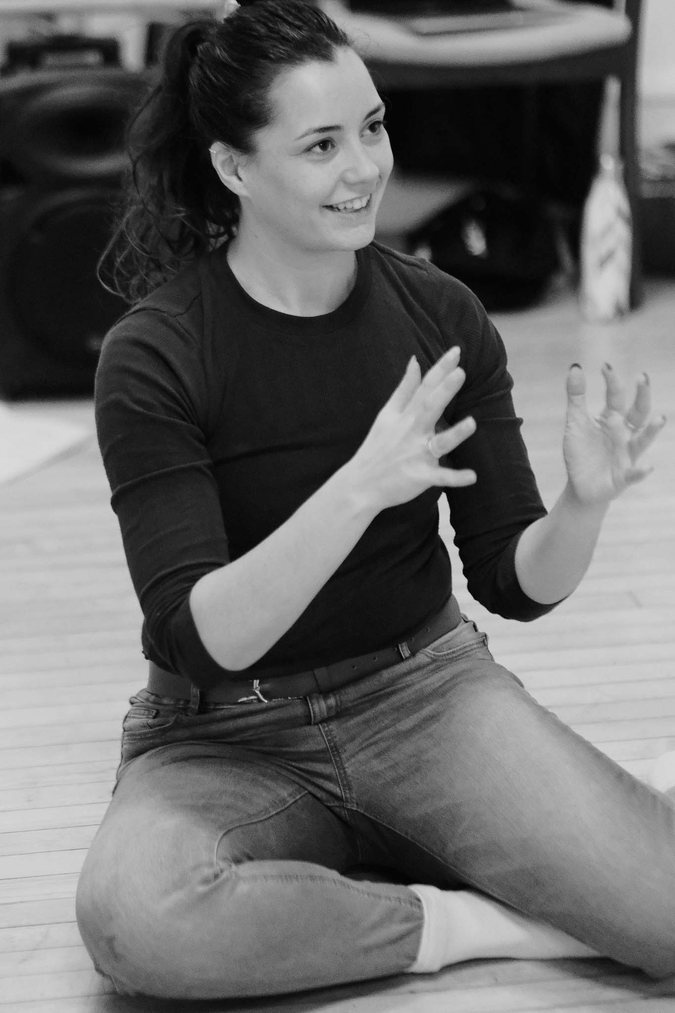 A picture of a young woman sat on the floor, smiling with her hands reaching out. It looks as though she is shaping something with her hands
