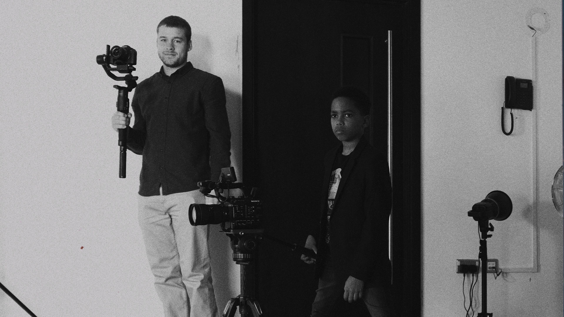 A film maker stands with a camera. Beside him is a young person with a camera on a tripod. The film maker is smiling at the camera while the young person is focused on filming something off screen