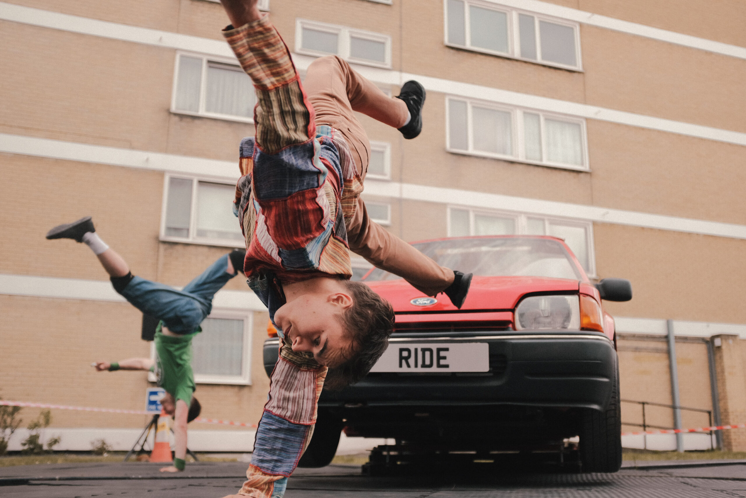 An image of a dance performance in the middle of a 60s looking housing estate in Southampton. In the middle of the performance space is a 1980s red car and the dancers are flipping off each side of it