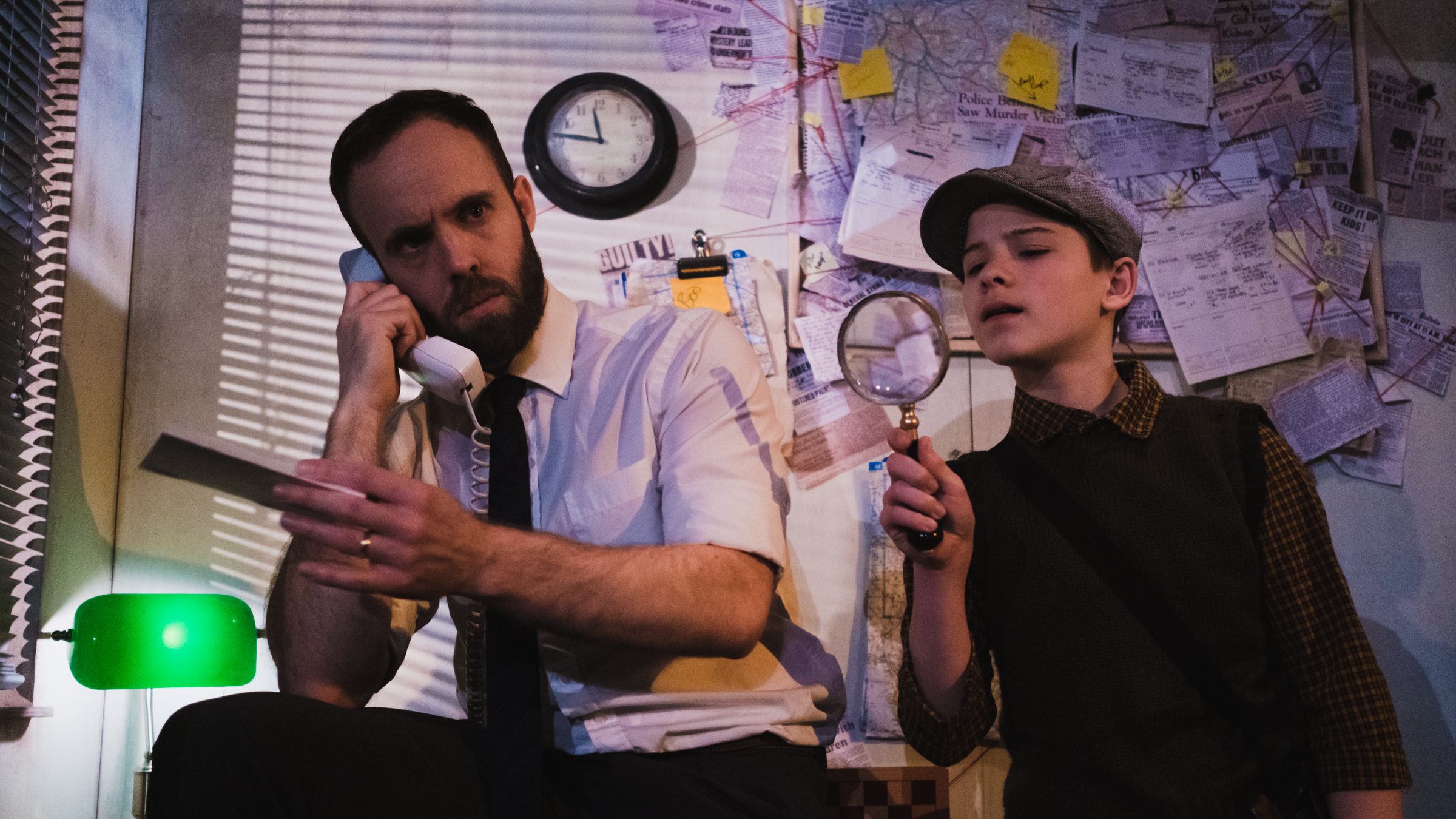 A dance theatre performance is taking place and it's set in the 1950s. It looks like something from film noir, there's a detective on the phone and a young boy next to him with a magnifying glass. The backdrop looks like a classic detective's office