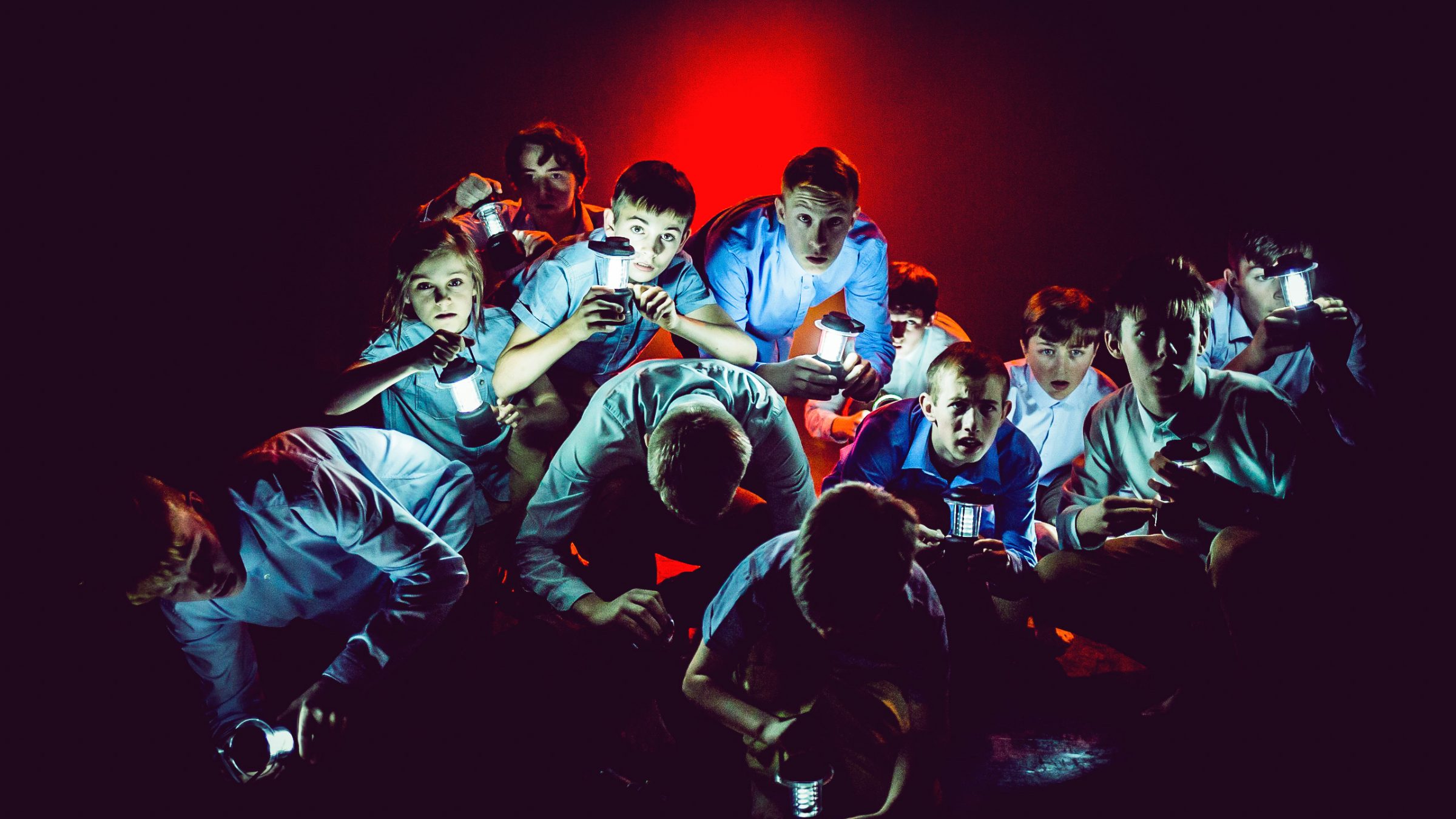 group of boys on a dark stage, shining torch lights in front of red back lighting. The boys are wearing blue tops and trousers and looking curious as they crouch in a huddle.