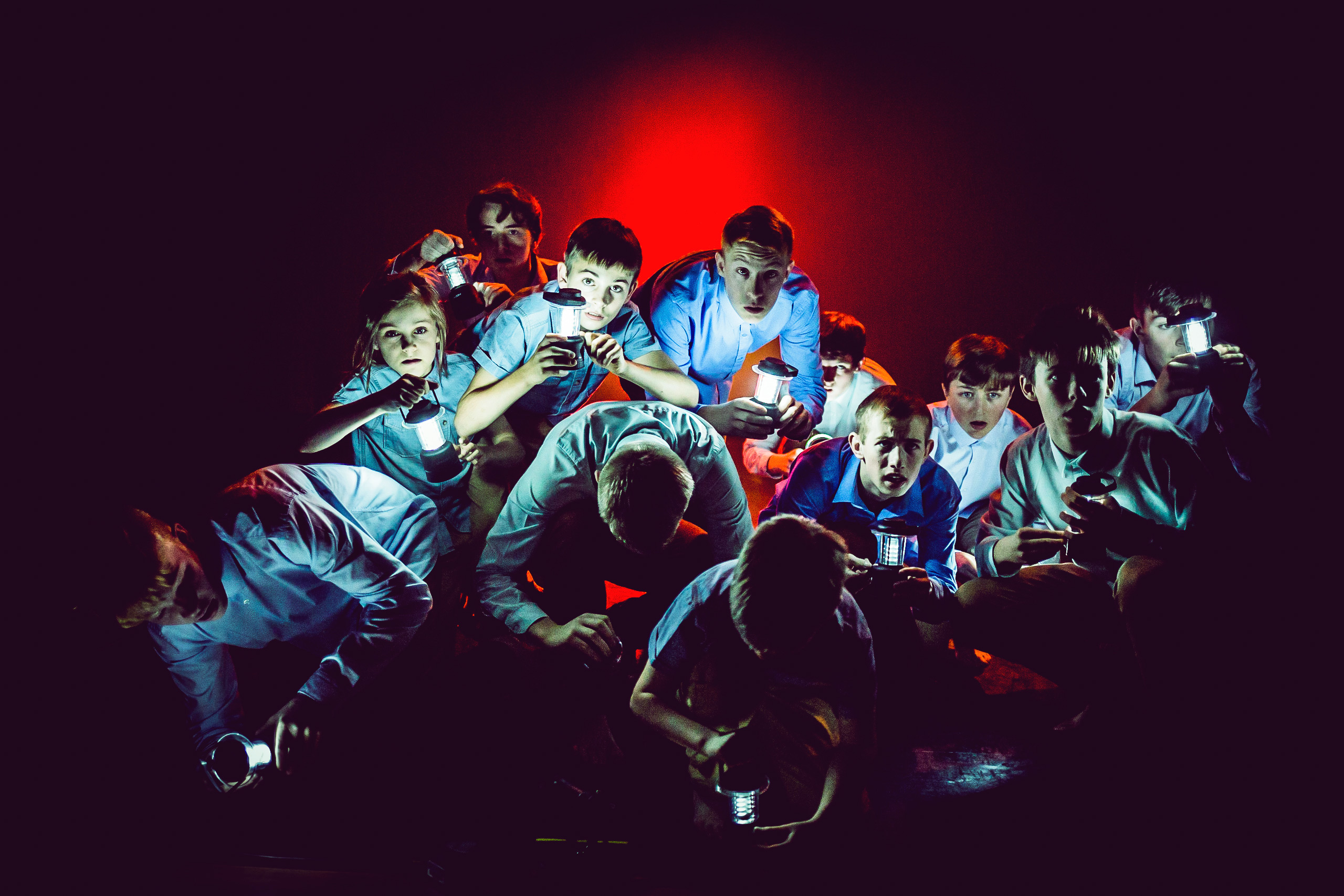group of boys on a dark stage, shining torch lights in front of red back lighting. The boys are wearing blue tops and trousers and looking curious as they crouch in a huddle.